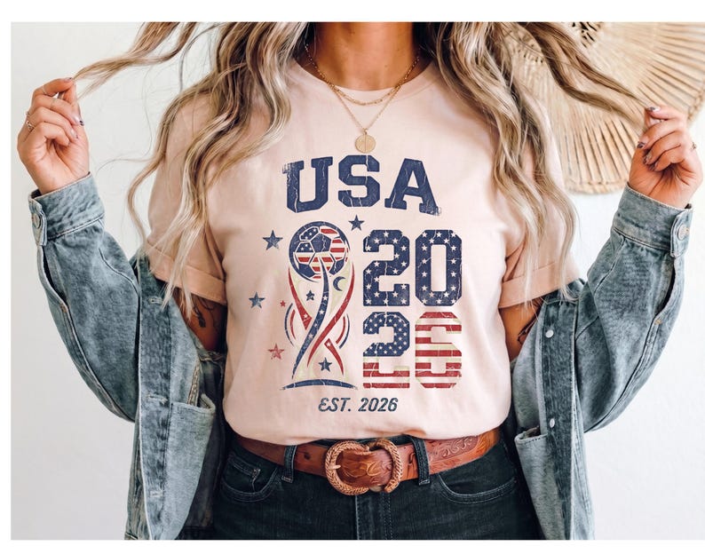 May include: A light peach-colored t-shirt featuring a distressed graphic with the text USA 2026 and EST. 2026. The design includes a soccer ball, stars, and stripes in red, white, and blue, layered over a stylized trophy shape with a worn texture.