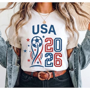 May include: A white short-sleeved t-shirt featuring a graphic design with the text USA 2026 in blue and red. The design includes a soccer ball atop a stylized trophy shape, surrounded by small red and blue stars, with the numbers 20 and 26.