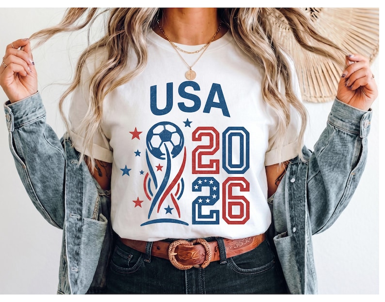 May include: A white short-sleeved t-shirt featuring a graphic design with the text USA 2026 in blue and red. The design includes a soccer ball atop a stylized trophy shape, surrounded by small red and blue stars, with the numbers 20 and 26.