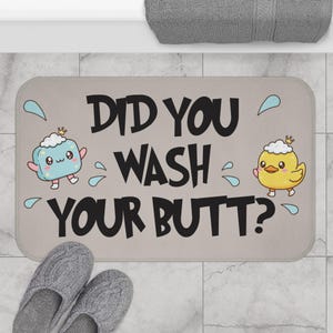 May include: A rectangular beige bath mat with the text "DID YOU WASH YOUR BUTT?" in black. The mat features cartoon illustrations of a soap bar and a chick, along with water droplet graphics.