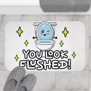 May include: White rectangular bath mat with a cartoon toilet illustration and the text "YOU LOOK FLUSHED!". The toilet has a smiling face and is surrounded by yellow starbursts. Two gray slippers are at the bottom.