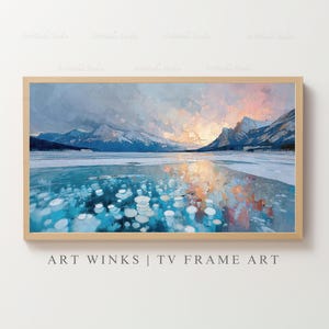 May include: A framed landscape painting depicts a serene winter scene. The artwork showcases a frozen lake with ice bubbles, snow-capped mountains, and a colorful sky. The frame is a light wood color. Text at the bottom reads: ART WINKS | TV FRAME ART.