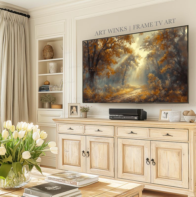 May include: A large framed TV displaying a landscape painting of a sunlit forest with golden and brown foliage. The TV is mounted above a light wood cabinet with drawers and doors. A vase of white tulips sits on a coffee table in front of the cabinet.