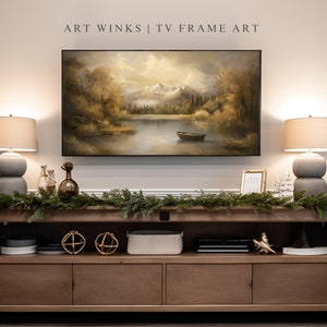 May include: A wooden media console with two drawers and two open shelves. The console is decorated with a garland of greenery, two gold orb decorations, and a gray speaker. A framed print of a lake scene with mountains in the background hangs above the console. Two lamps with gray bases and beige shades flank the print. The text "ART WINKS | TV FRAME ART" is printed above the print.