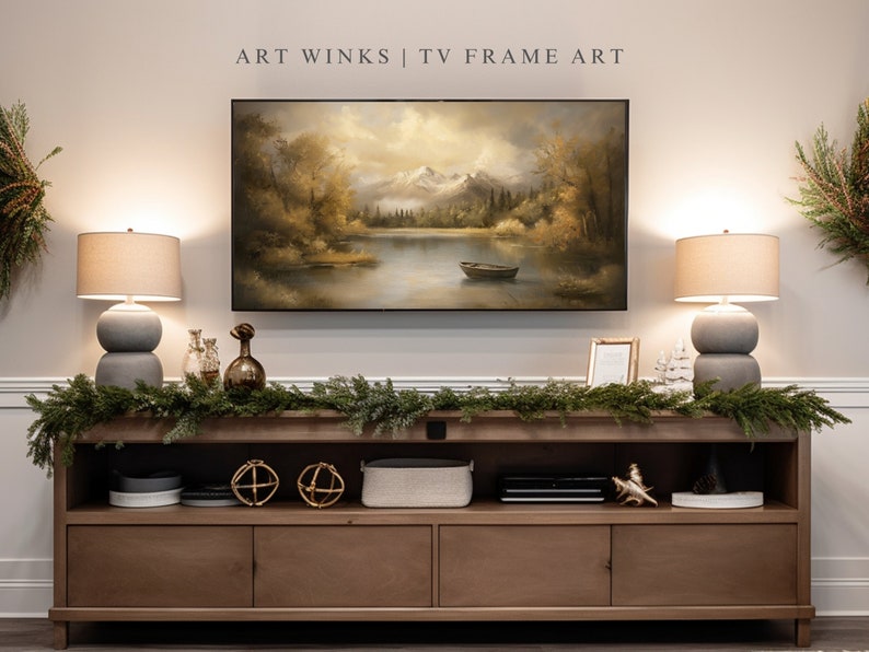 May include: A wooden media console with two drawers and two open shelves. The console is decorated with a garland of greenery, two gold orb decorations, and a gray speaker. A framed print of a lake scene with mountains in the background hangs above the console. Two lamps with gray bases and beige shades flank the print. The text "ART WINKS | TV FRAME ART" is printed above the print.