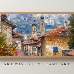 May include: A painting of a European village with a church steeple and mountains in the background. The buildings are painted in warm colors with a textured brushstroke style. The painting is framed in a light wood frame.