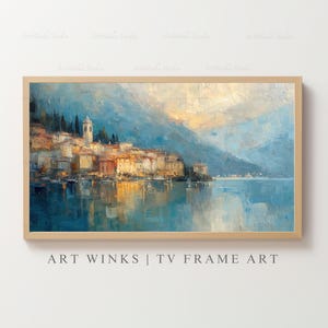 May include: A framed landscape painting depicts a coastal town with buildings in warm tones, a church tower, and a serene lake reflecting the sky. The artwork is set against a backdrop of blue mountains. The text "ART WINKS | TV FRAME ART" is at the bottom.