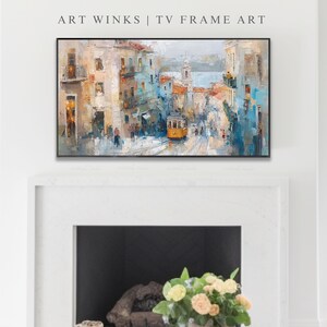Pu&ograve; includere: Una stampa artistica incorniciata intitolata "Art Winks | TV Frame Art" raffigura una scena di una citt&agrave; europea. Il dipinto presenta edifici nei toni del beige, blu e arancione, con un tram giallo su una strada acciottolata. L'opera &egrave; esposta sopra un camino.