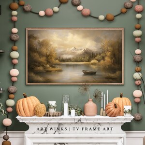 May include: A green wall with a framed painting of a lake and mountains. A string of decorative balls hangs on the wall. A fireplace mantel with two pumpkins and candles sits below the painting. The text "ART WINKS | TV FRAME ART" is on the mantel.