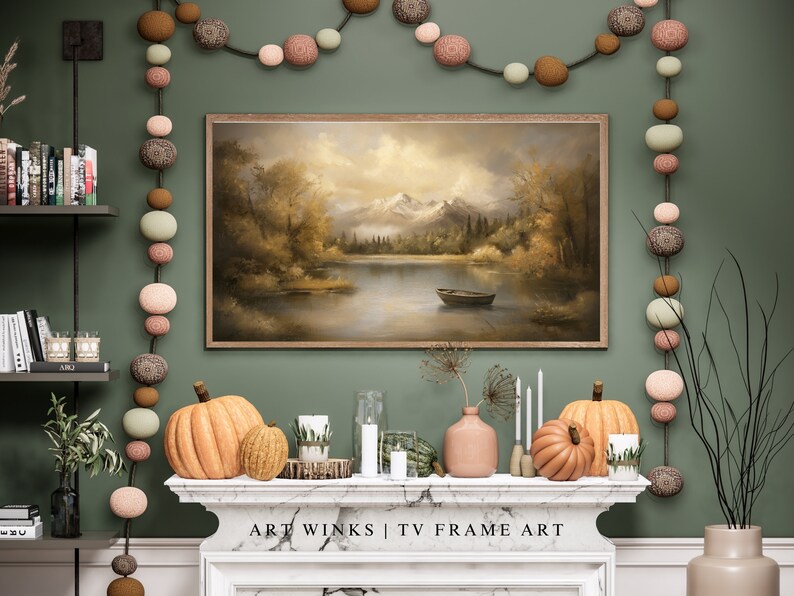 May include: A green wall with a framed painting of a lake and mountains. A string of decorative balls hangs on the wall. A fireplace mantel with two pumpkins and candles sits below the painting. The text "ART WINKS | TV FRAME ART" is on the mantel.