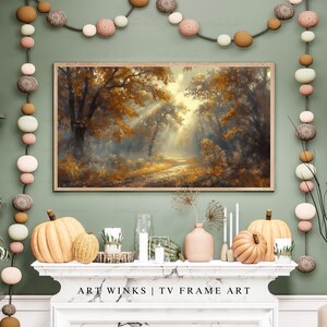May include: A framed landscape painting depicts a sunlit forest path with golden autumn foliage. The artwork is displayed above a fireplace decorated with pumpkins, candles, and decorative accents. The text "ART WINKS | TV FRAME ART" is visible.