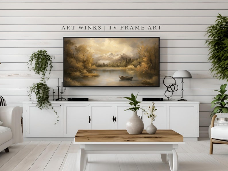 May include: A white living room with a white cabinet and a TV displaying a painting of a lake scene with mountains in the background. The TV is on a white cabinet with a black DVD player. There is a coffee table in front of the cabinet with a vase of flowers on it. There is a white couch on the left side of the image and a white chair on the right side of the image. The text "ART WINKS | TV FRAME ART" is displayed above the TV.