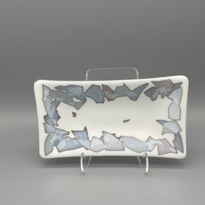 May include: A rectangular white dish with a decorative border of gray and blue geometric shapes. The dish is displayed on a clear acrylic stand, showcasing its design. The dish is likely used for serving or display.