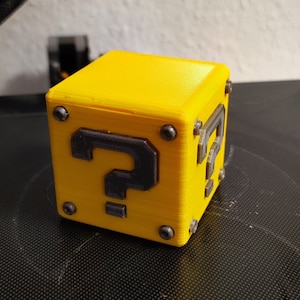 Puede incluir: Un bloque de interrogación amarillo impreso en 3D con un signo de interrogación negro y detalles plateados. El bloque está sobre una superficie negra.