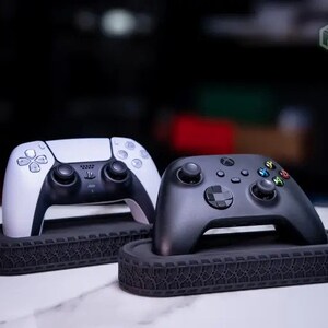 Könnte beinhalten: Zwei Gamecontroller, einer weiß und einer schwarz, ruhen auf strukturierten schwarzen Ständern. Der weiße Controller hat ein schlankes Design mit schwarzen Akzenten, während der schwarze Controller farbige Tasten aufweist. Die Ständer haben ein Reifenprofil-Design.