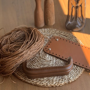 May include: A close-up of brown leather crafting supplies, including a spool of leather cord, two leather bag bottom pieces, and a leather handle. The supplies are arranged on a woven straw mat.