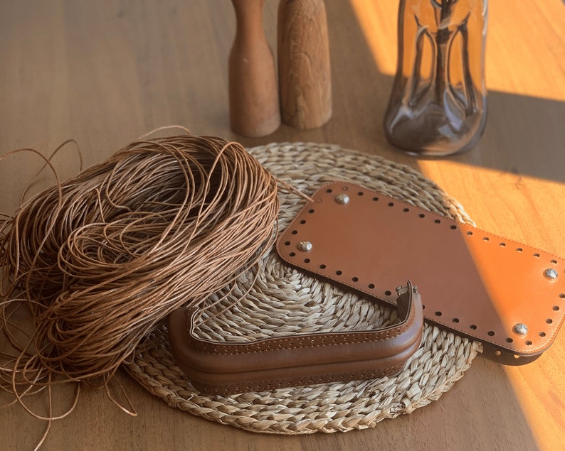 May include: A close-up of brown leather crafting supplies, including a spool of leather cord, two leather bag bottom pieces, and a leather handle. The supplies are arranged on a woven straw mat.