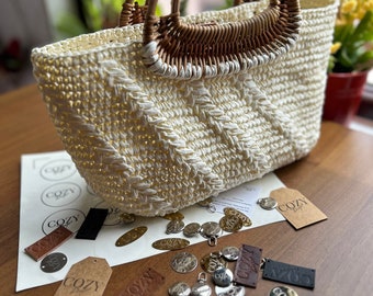 May include: A white crocheted tote bag with a woven brown handle. The bag is surrounded by various leather and metal tags with the word "Cozy" embossed on them.
