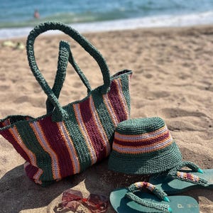 May include: A green and striped crochet beach bag, bucket hat, and sandals set on a sandy beach. The bag and hat feature burgundy, orange, and gray stripes. The sandals are green with matching crochet straps. Red sunglasses are also present.