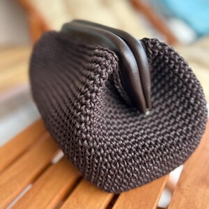 May include: A brown crocheted bag with a leather top closure. The bag is sitting on a wooden surface.