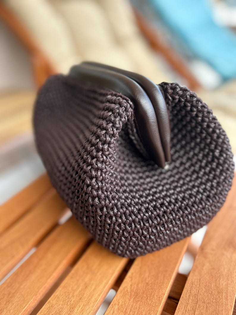 May include: A brown crocheted bag with a leather top closure. The bag is sitting on a wooden surface.