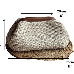 May include: A cream-colored crocheted clutch bag with a brown leather top handle. The bag measures 8 inches wide, 7 inches tall, and 11 inches long.