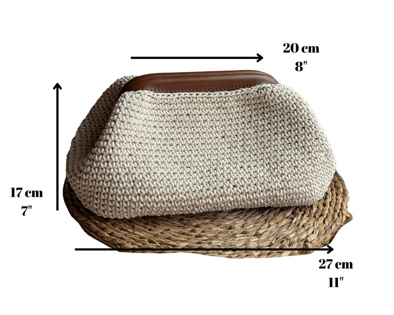 May include: A cream-colored crocheted clutch bag with a brown leather top handle. The bag measures 8 inches wide, 7 inches tall, and 11 inches long.