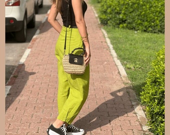 May include: A woman wearing a black tank top and lime green pants with a black and brown woven bag slung over her shoulder. She is walking on a brick pathway.