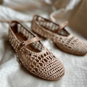 May include: A pair of beige woven flats with a Mary Jane strap and buckle closure. The shoes have a textured, open weave design and a light brown leather strap. The shoes are set against a soft, neutral background.