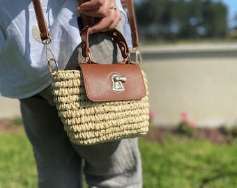May include: A small straw handbag with a brown leather flap and a silver lock closure. The bag has a top handle and a shoulder strap.