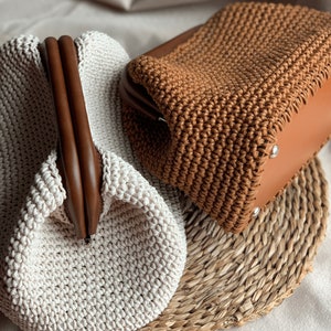 May include: Two crocheted handbags, one in beige and one in brown, with wooden handles. The handbags are sitting on a woven straw mat.