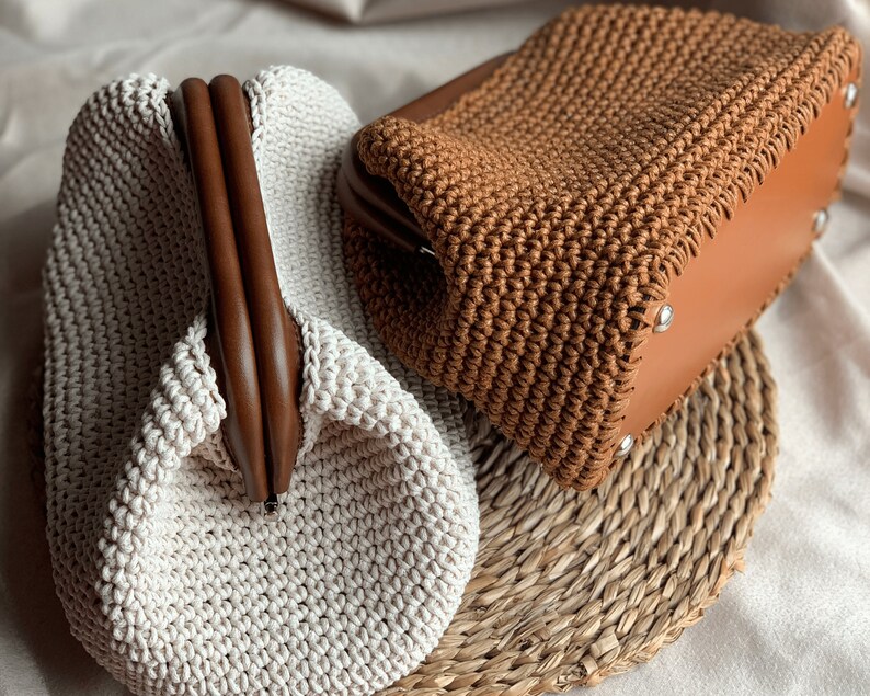 May include: Two crocheted handbags, one in beige and one in brown, with wooden handles. The handbags are sitting on a woven straw mat.