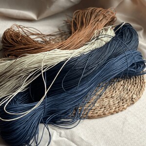 May include: Three spools of cord in different colors: brown, white, and dark blue. The cords are wound around each other and are sitting on a woven mat.
