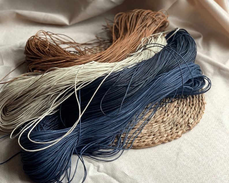 May include: Three spools of cord in different colors: brown, white, and dark blue. The cords are wound around each other and are sitting on a woven mat.