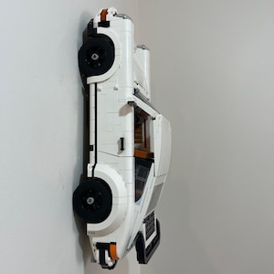 May include: A white plastic brick-built model car featuring black wheels, orange side marker lights, and a visible interior with brown seating. The vehicle is constructed from interlocking building blocks and is displayed in a side profile view.