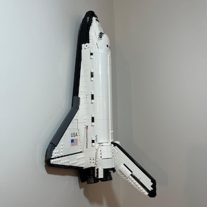 May include: A white and black space shuttle model constructed from building blocks. The fuselage features the text United States, the NASA logo, and a small American flag decal. The shuttle is mounted vertically, displaying its wings and engine area.