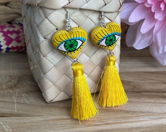 May include: A pair of yellow heart-shaped earrings with green eyes and yellow tassels. The earrings are hanging from a woven basket.