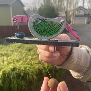 May include: A handcrafted wooden bird toy with a pink head and tail, gray body with green wing details, and a small blue nest. A string connects the bird to a small brown ball, creating a playful design.