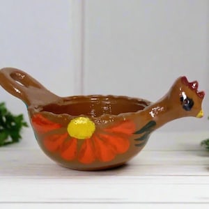 Pode incluir: Uma tigela de cerâmica marrom em forma de galinha, com uma alça e um desenho floral. A tigela tem uma crista vermelha, um olho preto e um bico amarelo. A flor tem pétalas laranja e um centro amarelo.