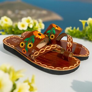 May include: A pair of brown leather sandals with embroidered sunflowers and green leaves. The sandals have a toe thong and a thick sole.