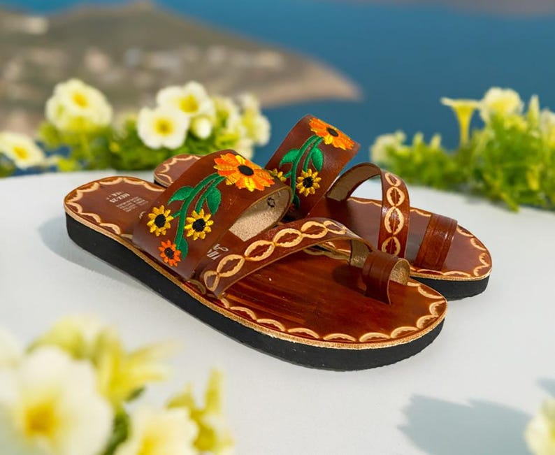 May include: A pair of brown leather sandals with embroidered sunflowers and green leaves. The sandals have a toe thong and a thick sole.