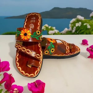 May include: Brown leather sandals with a toe loop and embroidered sunflowers. The sandals have a brown leather strap with a braided design.