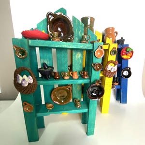 Può includere: Un'esposizione di miniature di ceramica per la cucina, tra cui ciotole, tazze e cestini, disposte su una mensola di legno verde acqua, gialla e blu. La ceramica è nei toni del marrone, arancione e nero, con un pezzo decorativo rosso in cima.