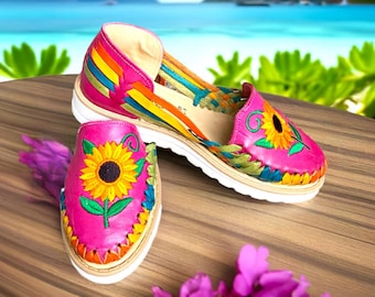 May include: A pair of pink leather sandals with colorful woven straps and embroidered sunflowers. The sandals have a white platform sole.