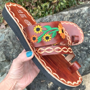 May include: Brown leather sandals with a black sole and embroidered sunflowers. The sandals have a toe loop and two straps.