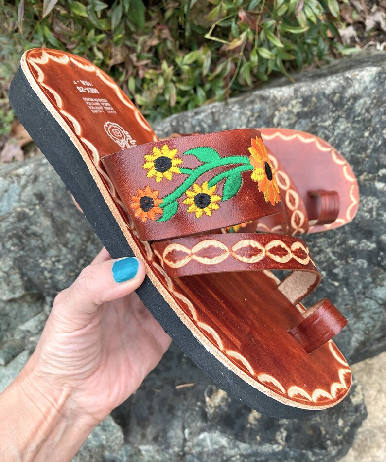 May include: Brown leather sandals with a black sole and embroidered sunflowers. The sandals have a toe loop and two straps.