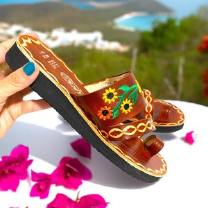 May include: Brown leather sandals with a floral embroidery design featuring sunflowers and green leaves. The sandals have a toe loop and a thick sole.