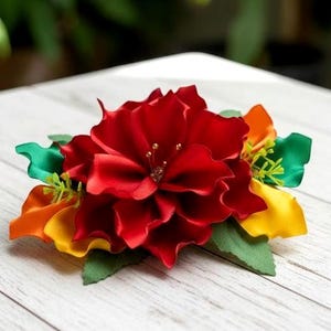 May include: A vibrant, handmade floral hair accessory. The centerpiece is a large, layered red flower, accented with green, orange, and yellow petals. The flower is set against a light wooden surface, creating a striking contrast.
