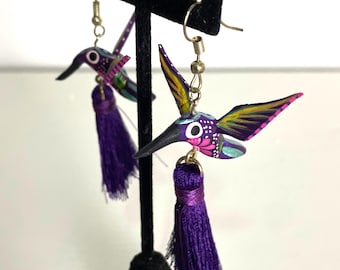 May include: A pair of purple hummingbird earrings with gold hooks and purple tassels. The hummingbirds are painted with bright colors and intricate details.