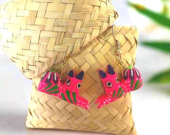 May include: A pair of handmade, vibrant pink and green wooden earrings. The earrings feature a stylized bird design with black accents and gold-colored ear wires. They are displayed on a woven straw background.